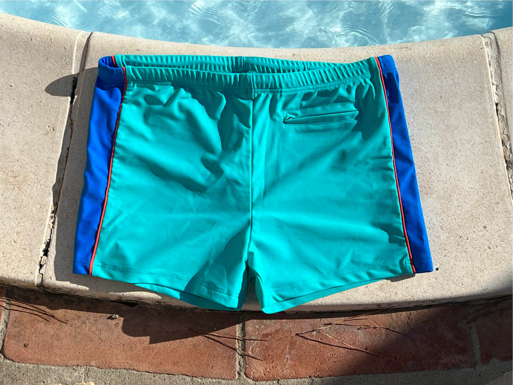 Pair of swimtrunks in mintish green and cobalt blue side lying flat on the ground at a swimming pool