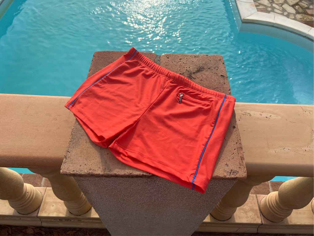 Swimtrunk in orange red with cobalt blue contrast lying flat on a pole at a swimming pool